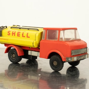 May include: A vintage toy Shell tanker truck. The truck features a bright yellow tank with the Shell logo and red cab. The wheels are black, and the truck is set against a neutral background.