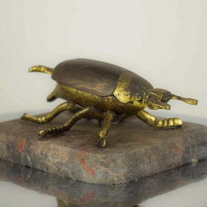 May include: A decorative brass beetle figurine with a dark brown shell and gold-colored legs and accents. The beetle is perched on a rectangular, mottled stone base with red and gray hues. The figurine has a detailed, antique appearance.
