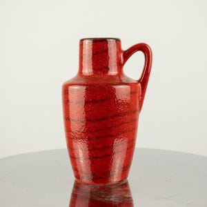 May include: A red ceramic vase with a handle. The vase has a textured surface with dark brown horizontal stripes. The vase is approximately 8 inches tall.