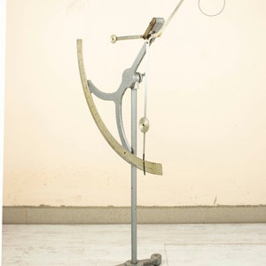 May include: A silver-colored, freestanding, scientific instrument with a graduated arc and a hanging weight. The instrument is used to measure the specific gravity of liquids.
