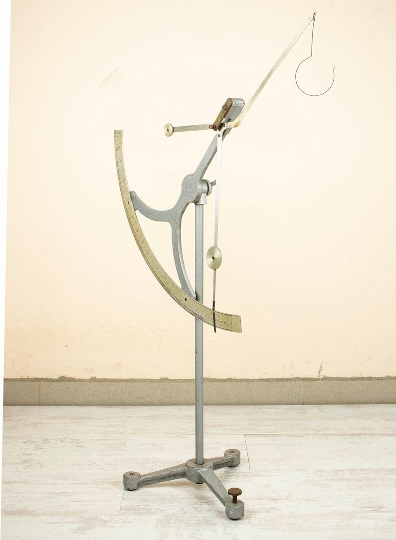 May include: A silver-colored, freestanding, scientific instrument with a graduated arc and a hanging weight. The instrument is used to measure the specific gravity of liquids.