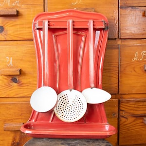 Può includere: Un portautensili da cucina da parete in metallo smaltato rosso con un vassoio raccogligocce inferiore e una barra orizzontale che sostiene tre utensili appesi. Il set include un mestolo rotondo, una schiumarola forata e una spatola.