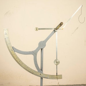 May include: A silver and white metal scale with a circular protractor and a hanging hook. The scale is on a white background.