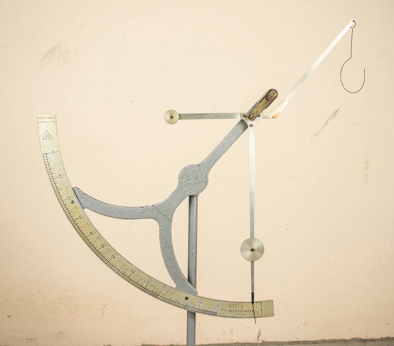 May include: A silver and white metal scale with a circular protractor and a hanging hook. The scale is on a white background.