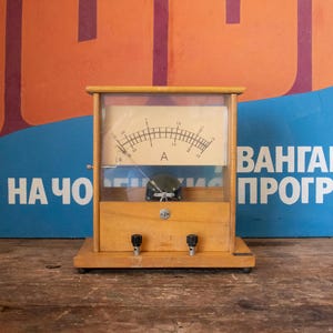 Può includere: Un amperometro analogico vintage in legno con un quadrante in vetro e un ago nero. L'amperometro è etichettato con "A" e ha segni da 0 a 5. L'amperometro è seduto su una base in legno con due manopole nere.