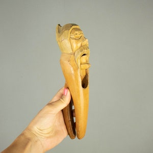 Op de afbeelding: Een houten notenkraker gebeeldhouwd in de vorm van een mannenhoofd met een baard. De notenkraker is geschilderd in tinten bruin en heeft een scharnierende kaak.