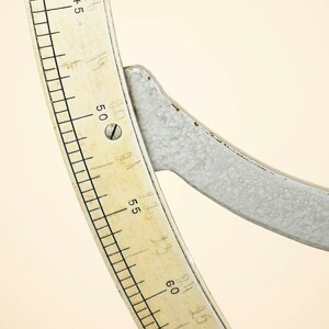 May include: Close-up of a white metal protractor with a cream-colored scale marked in degrees. The scale is numbered from 45 to 65 degrees.