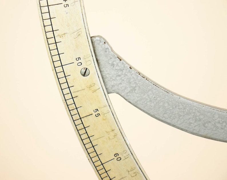 May include: Close-up of a white metal protractor with a cream-colored scale marked in degrees. The scale is numbered from 45 to 65 degrees.