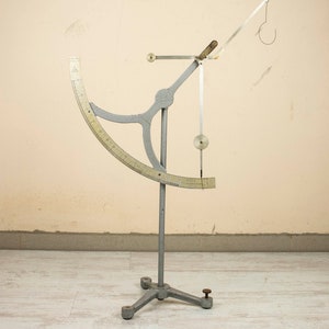 May include: A silver-colored scientific instrument with a graduated arc and a hanging weight. The instrument is mounted on a tripod stand.