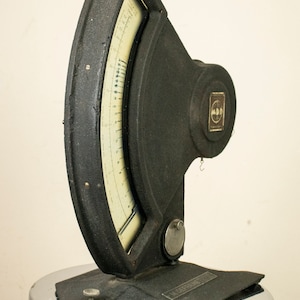 May include: A black vintage scale with a circular dial and a metal base. The scale has a white dial with black markings and a needle that points to the weight. The scale is labeled "M.D.D. Kalibrator".