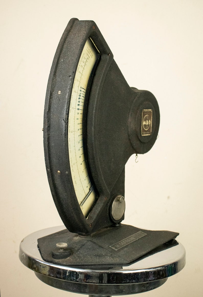 May include: A black vintage scale with a circular dial and a metal base. The scale has a white dial with black markings and a needle that points to the weight. The scale is labeled "M.D.D. Kalibrator".