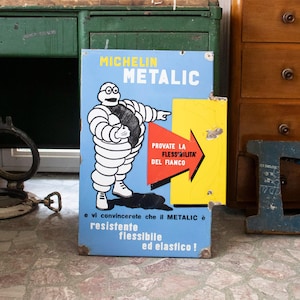 May include: Vintage Michelin Metalic sign with the Michelin Man, a white figure made of tires, pointing to a yellow arrow. The sign is blue with Italian text and a red arrow. The sign shows signs of wear.