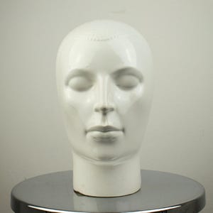 Puede incluir: Una escultura de cabeza de cerámica blanca con los ojos cerrados. La cabeza es lisa y brillante, con rasgos faciales sutiles y un patrón texturizado en la parte superior. Se exhibe sobre una superficie metálica reflectante.