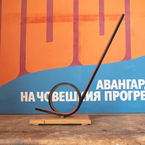 Op de afbeelding: Een zwart metalen sculptuur met een rode streep, gevormd als een lange, dunne S, met een ronde lus aan de onderkant. Het sculptuur is gemonteerd op een houten basis. Het sculptuur staat voor een blauwe en oranje poster met de tekst "АВАНГАРД НА ЧОВЕШКИЯ ПРОГРЕС".