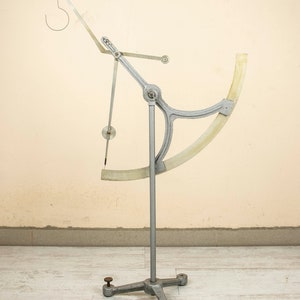 May include: A silver metal scientific instrument with a white plastic protractor and a hook at the top. The instrument is mounted on a tripod stand.