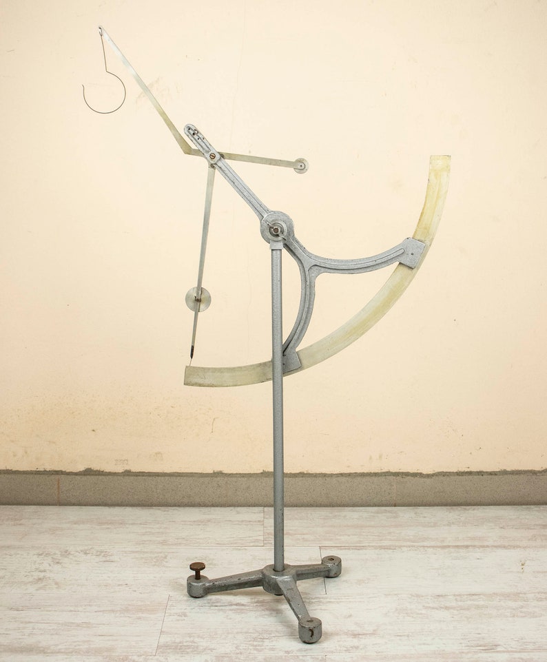 May include: A silver metal scientific instrument with a white plastic protractor and a hook at the top. The instrument is mounted on a tripod stand.