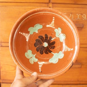 Könnte beinhalten: Eine runde Terrakottaschale mit einem dunkelbraunen Blumenmotiv in der Mitte, umgeben von vier hellgrünen floralen Akzenten und weißen, gepunkteten Linienmustern, die zum Rand hin verlaufen. Die Innenfläche ist glatt mit einem rotbraunen Finish.