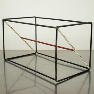 May include: A rectangular wire frame sculpture constructed from thin black metal rods. The structure features a single diagonal white rod on the side and a horizontal red rod connecting the centre of the frame, showing signs of paint wear.