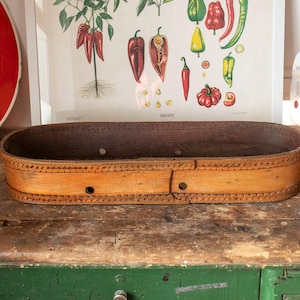 Può includere: Cesto ovale in legno con esterno marrone chiaro e interno marrone scuro. Il cesto presenta cuciture decorative sul bordo ed è appoggiato su una superficie in legno rustico. Un cartello vintage e una stampa botanica sono sullo sfondo.