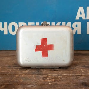 May include: A vintage metal first aid kit with a red cross painted on the front. The kit has a silver finish and a metal latch.