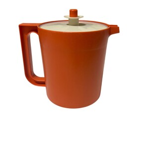 May include: An orange plastic pitcher with a handle and a lid. The pitcher has a cylindrical body and a spout. The lid is off-white with an orange handle. The pitcher is likely vintage and suitable for serving beverages.