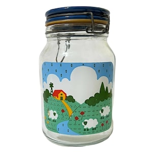 May include: A glass jar with a blue lid and a whimsical illustration of a farm scene with a red house, green trees, and white sheep in a field. The jar has a blue and white checkered pattern around the top.