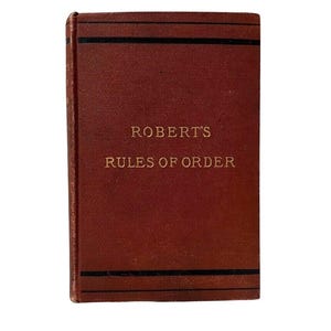 May include: A hardcover book with a dark red cover. The title "ROBERT'S RULES OF ORDER" is printed in gold lettering. Two black horizontal lines are at the top and bottom of the cover. The book is a classic guide to parliamentary procedure.