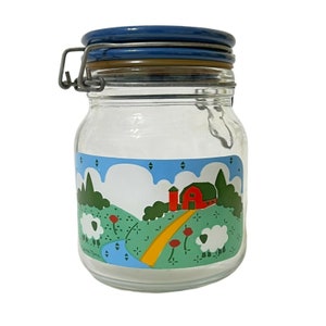 May include: A glass jar with a blue lid and a farmhouse scene with a red barn, green trees, white sheep, and yellow path. The jar has a metal latch.