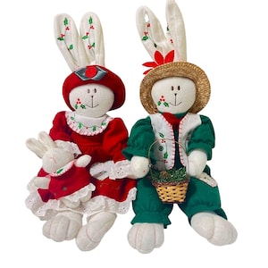 May include: A set of three plush bunny Christmas decorations. The two larger bunnies are sitting and wearing festive clothing. The female bunny is wearing a red dress with white lace trim and a red hat. The male bunny is wearing a green vest and pants with a brown basket. The smaller bunny is wearing a red dress and is sitting on the female bunny's lap.