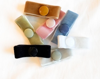 May include: A collection of velvet fabric button closures in various colors, including tan, green, pink, blue, black, and white. Each closure features a matching round button. The items are arranged on a white background.