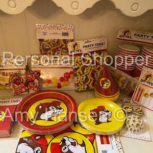 May include: A Buc-ee's themed birthday party supply set with red and yellow plates, cups, napkins, a party hat, balloons, candles, and cupcake liners. The set features the Buc-ee's mascot, a beaver wearing a red cap.