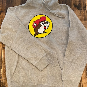May include: A gray hooded sweatshirt with a yellow circle logo featuring a cartoon beaver wearing a red hat. The logo says "Beaver"