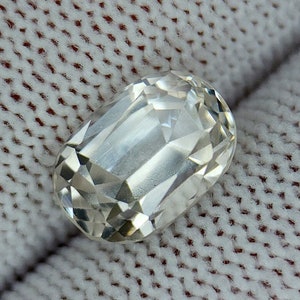 May include: A clear, faceted, oval-shaped gemstone with a slightly yellowish hue. The gemstone is resting on a white, textured surface.