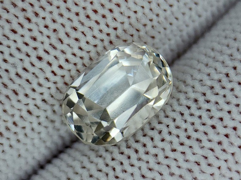 May include: A clear, faceted, oval-shaped gemstone with a slightly yellowish hue. The gemstone is resting on a white, textured surface.