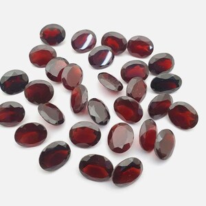 May include: A collection of oval-cut, deep red gemstones scattered on a white surface. The gems have a polished, faceted appearance, reflecting light and showcasing their rich color. These gemstones are ideal for jewelry making.