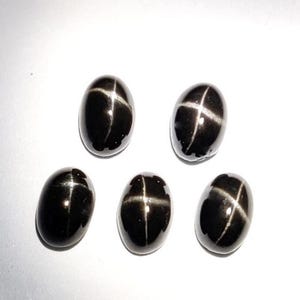 以下が含まれることがあります： 磨かれた楕円形の黒いスターサファイアが5つ。各宝石は、表面に目立つ白い四条の星模様があります。石は白い背景に配置され、暗い色と反射特性を強調しています。