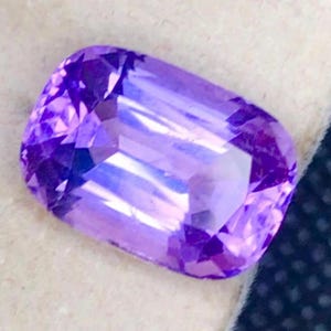 Könnte beinhalten: Ein rechteckig geschliffener Edelstein in einem tiefen Violettton. Die Oberfläche des Edelsteins ist facettiert und reflektiert das Licht, wodurch seine innere Struktur sichtbar wird. Helle Schattierungen sind sichtbar.