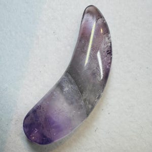 May include: A crescent-shaped amethyst crystal, polished smooth and featuring a light purple hue with darker streaks throughout.