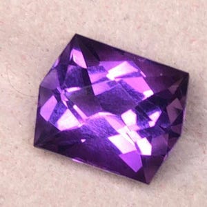May include: A faceted, deep purple gemstone with a geometric cut. The gem has multiple reflective surfaces that catch the light, creating a sparkling effect. The gemstone is set against a neutral background.