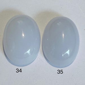 May include: Two oval, pale blue gemstones with a smooth, polished, and glossy surface. Each stone has a number printed on it: 34 and 35. The gemstones are set against a white background. Ideal for jewellery making.