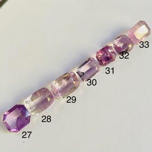 May include: Eight faceted gemstones in shades of purple and pink, arranged diagonally. Each gem has a rectangular shape with cut corners. Numbers 27 to 33 are visible next to each gem. The gems are displayed on a light-coloured surface.