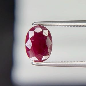 Peut inclure: Une pierre précieuse de rubis ovale et facettée, tenue par une pince. Le rubis est d'un rouge profond et riche, avec de multiples facettes qui réfléchissent la lumière. La pince est de couleur argentée et maintient la pierre sur un fond flou.