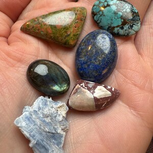 Op de afbeelding: Een verzameling gepolijste edelstenen in verschillende vormen en kleuren. Inclusief een turkoois steen, een blauwe lapis lazuli, een groene en rode unakiet, een labradoriet, een rood-witte jaspis en een blauwe kyaniet.