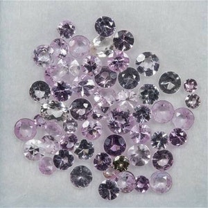 Puede incluir: Una colección de piedras preciosas sueltas de color rosa y morado, probablemente zafiros, en varios tamaños y formas. Las piedras preciosas están dispuestas sobre una superficie blanca.