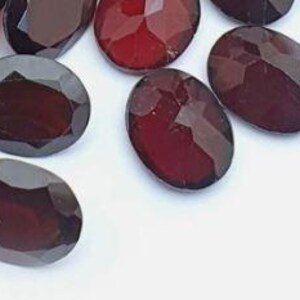 May include: Close-up of several oval-cut, dark red gemstones. The gems have a faceted surface, catching the light and displaying a deep, rich color. The gemstones are scattered on a white surface.