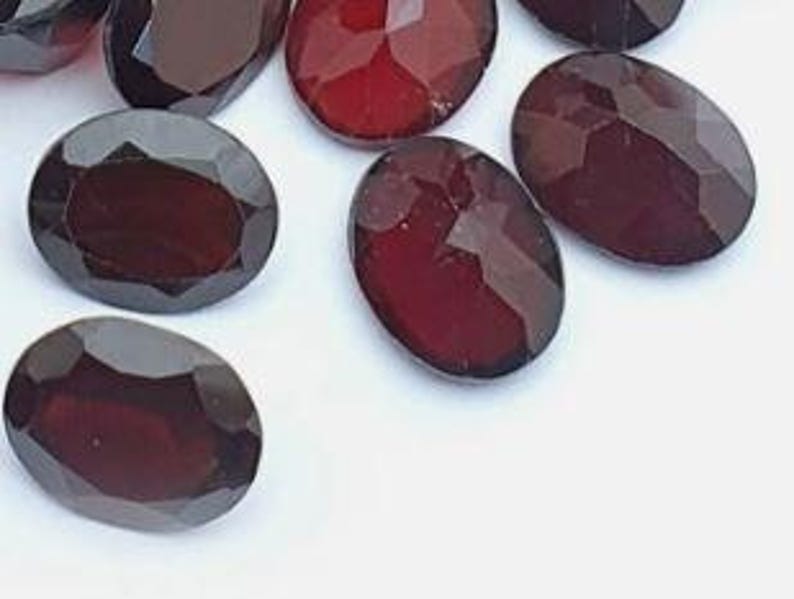May include: Close-up of several oval-cut, dark red gemstones. The gems have a faceted surface, catching the light and displaying a deep, rich color. The gemstones are scattered on a white surface.