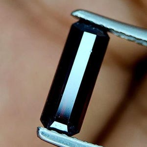 May include: A dark, rectangular gemstone with a polished surface is held by silver tweezers. The gemstone has a long, slender shape with a bright, white reflection running down its centre.