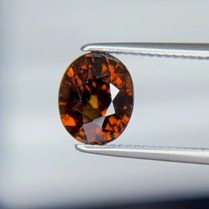 May include: An oval-shaped gemstone held by tweezers. The gem displays a blend of dark brown and orange colours, with facets that reflect light. The gemstone's form is elongated, and the cut reveals internal reflections.