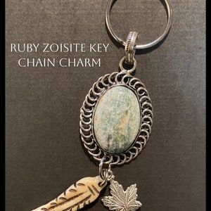 May include: A silver key chain charm featuring a ruby zoisite stone in an oval frame. It includes a carved bone feather and a silver maple leaf charm. The image text reads "RUBY ZOISITE KEY CHAIN CHARM".