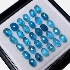 May include: A collection of oval-shaped, bright blue gemstones arranged in rows within a black display case. The gemstones have a polished, translucent appearance, showcasing internal patterns and variations in color.